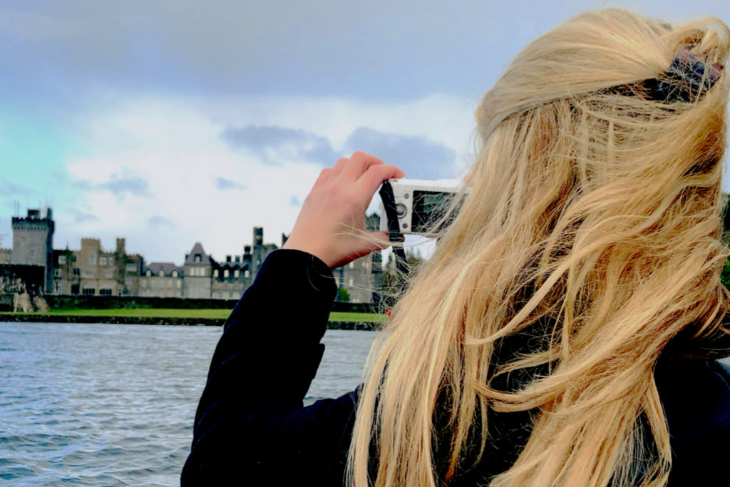 A blonde woman takes photos of Ashford Castle from a hand carved boat.